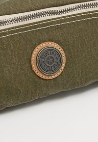 Sac en tissu vert avec une fermeture éclair argentée et un patch rond en métal portant le logo Kipling, cousu avec un fil beige.