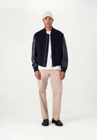 Laivastonsininen bomber-takki nahkaisilla hihoilla, vetoketjulla edessä ja resorireunuksilla, valkoisen T-paidan päällä, yhdistetty beigen värisiin housuihin ja mustiin kenkiin.