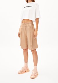Cropped white t-shirt with black text, beige high-waisted shorts with tied belt, worn with light pink sneakers and matching socks.