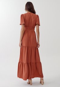 Maxiklänning i terrakottafärgad tyg med texturerade lager, puffärmar och åtsittande midja. Har en diskret bakre dragkedja och utsvängd silhuett.