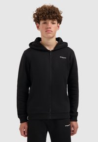 Jongere met krullend haar, gekleed in een zwarte rits-hoodie en bijpassende broek met kleine witte "BALLIN'" logo's, staand tegen een grijze achtergrond.