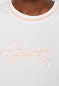 Närbild av en vit t-shirt med en ljusrosa krage och "Guess" broderat över bröstet i matchande rosa tråd.