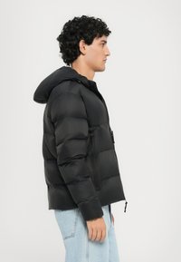 Schwarze Daunenjacke mit Kapuze, die horizontale Nähte, eine Reißverschlusstasche und einen gerafften Saum aufweist. Das Modell trägt hellblaue Jeans.