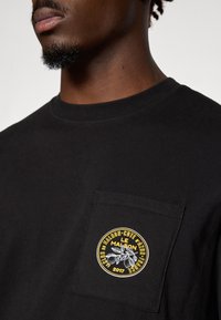 Svart bomullst-skjorte med en rund, brodert lapp på venstre lomme med et bladdesign og tekst i gult og hvitt.