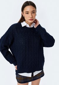 Pullover navy in maglia a trecce con maniche lunghe, polsini e orlo a coste. Abbinato a una camicia bianca con colletto e una gonna mini a scacchi.