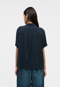 Γυναίκα με μακριά σκούρα μαλλιά φορώντας μπλε σκούρο μπλουζάκι με κοντά μανίκια και μπλε τζιν παντελόνι, όρθια με την πλάτη προς τα εμπρός σε απλό φόντο.