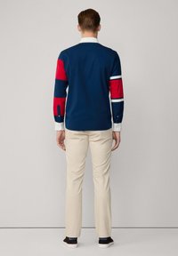 Camisa de manga larga azul con acentos a rayas rojas y blancas en las mangas, combinada con pantalones beiges y zapatos oscuros. Diseño sencillo, material de algodón.