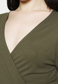 Haut enveloppant vert olive en tissu doux, présentant un décolleté en V profond et des détails brodés, avec un léger drapé sur l'avant.