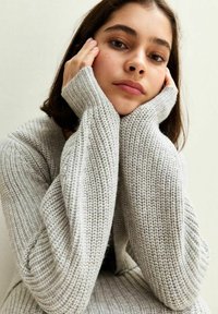 Pull en maille gris clair avec des poignets côtelés et une texture, présentant de longues manches et une coupe ample, porté par un modèle avec les mains reposant sur le visage.