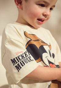 Cremefarvet bomulds T-shirt med et stort Mickey Mouse print i brune og sorte nuancer, med teksten "Disney Mickey Mouse" i fed skrift på ærmet.