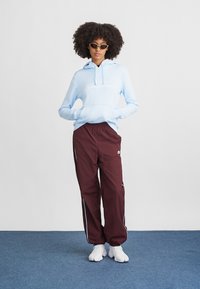 Svetlomodrá mikina s kapucňou a bielou grafikou, spárovaná s bordovými teplákmi so stranovými pruhmi. Biele športové topánky dopĺňajú vzhľad.