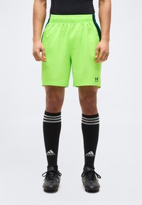 Άνδρας που φοράει neon πράσινα αθλητικά σορτς Under Armour, μαύρες κάλτσες Adidas μέχρι το γόνατο και μαύρες ποδοσφαιρικές παπούτσια, στέκεται μπροστά σε απλό φόντο.