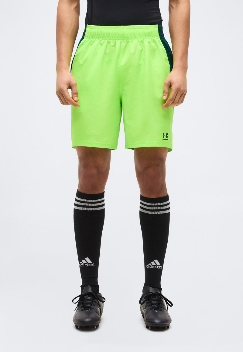 Άνδρας που φοράει neon πράσινα αθλητικά σορτς Under Armour, μαύρες κάλτσες Adidas μέχρι το γόνατο και μαύρες ποδοσφαιρικές παπούτσια, στέκεται μπροστά σε απλό φόντο.