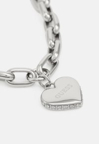 Silver chain bracelet featuring a heart charm with "GUESS" engraved and bordered by clear rhinestones. Smooth and shiny metal finish.