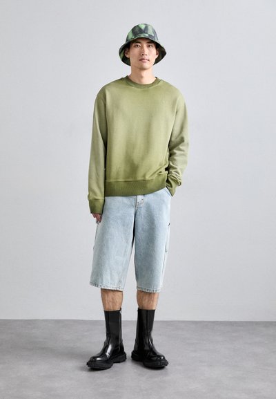 Felpa verde, pantaloni cargo azzurri chiari e stivali neri con zip. Il modello indossa un cappello a secchiello decorato. Sfondo in cemento.