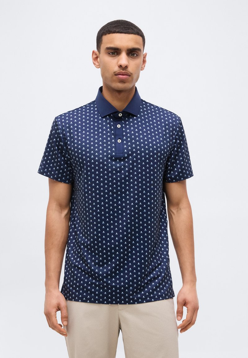 Άνδρας με κοντά μαλλιά, φορώντας μπλε ναυτικό πόλο με μικρά λευκά σχέδια και μπεζ παντελόνι, στέκεται μπροστά σε ένα απλό ανοιχτό φόντο.