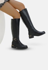 Bottes noires montantes en matériau synthétique lisse, avec un petit talon, des fermetures éclair sur le côté et une sangle décorative avec un accent doré.