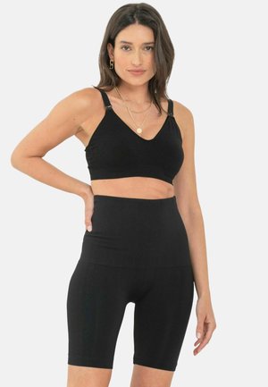 Kobieta w czarnym sportowym biustonoszu i wysokich szortach rowerowych, pozująca z jedną ręką na biodrze na jednolitym tle.