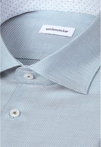 Chemise habillée texturée bleu clair pour homme avec des boutons blancs et une étiquette blanche portant la mention "seidensticker" à l'intérieur du col.