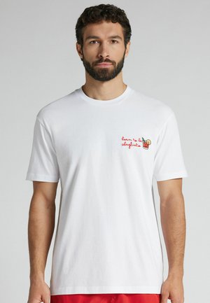 Man met baard draagt een wit T-shirt met rode tekst "born to be sbagliato" en een kleine cocktailafbeelding op de borst, staand tegen een effen achtergrond.