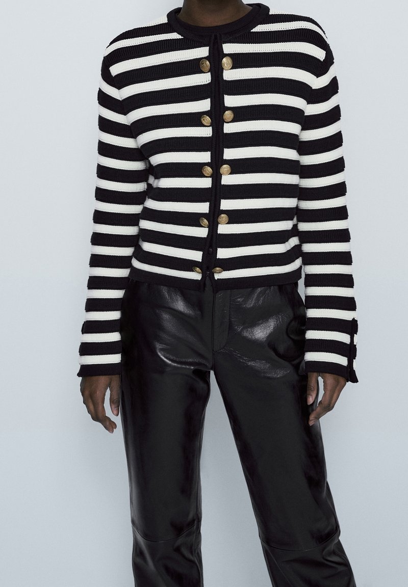 Cardigan à rayures noir et blanc avec une texture tricotée, doté de boutons dorés et d'un col rond, associé à un pantalon en cuir noir brillant.