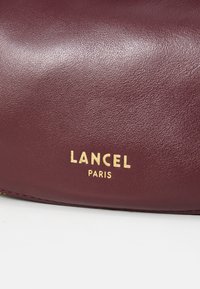 Sac en cuir bordeaux avec une texture lisse et le logo doré "LANCEL PARIS" embossé à l'avant. Design simple et discret.