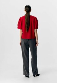 Haut rouge à manches courtes avec des manches bouffantes et des découpes élaborées, associé à un pantalon gris taillé et des escarpins noirs.