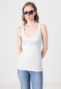 Ljust grön ribbad linne med rundad halsringning och silverfärgad "GUESS"-logotyp. Bärs med mörka solglasögon och blå jeans. Slät textur.