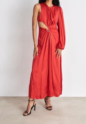 Femme en robe longue rouge à une manche longue, découpe latérale, détail anneau circulaire à la taille, et sandales à talons hauts noires.