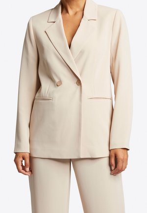 Kobieta ubrana w beżową marynarkę dwurzędową z wyciętymi klapami i dopasowane spodnie, stojąca z rękami swobodnie opuszczonymi wzdłuż ciała.