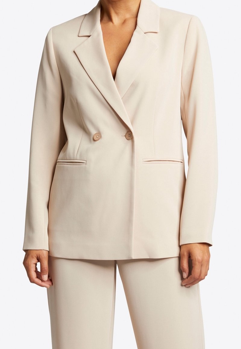 Femme portant un blazer beige croisé à revers crantés et un pantalon assorti, debout les mains détendues le long du corps.