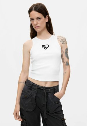 Jonge vrouw met lang bruin haar die een wit mouwloos cropped topje draagt met een zwart hartmotief en zwarte cargobroeken met zakken.