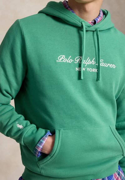Άτομο που φοράει πράσινο φούτερ Polo Ralph Lauren με λογότυπο και καρό πουκάμισο από μέσα, με το χέρι στην μπροστινή τσέπη.