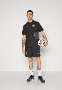 Sort polo med gule detaljer og logo, parret med sorte shorts. Holder en fodbold, iført sorte træningssko og hvide sokker.