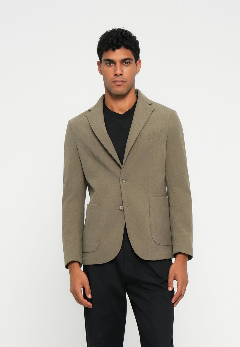 Grijs-groene blazer in een gestructureerde stof, met een notch-lapel, twee voorzakken en een sluiting met één knoop, gecombineerd met een zwart shirt.