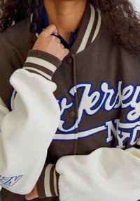 Brązowa kurtka typu varsity z białymi rękawami. Ma niebieski haftowany napis i prążkowane mankiety. Z przodu widoczne są napy.