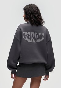 Übergroßer grauer Sweatshirt mit glatter Textur und dem Wort "BERLIN" in einer gewellten, hellgrauen Schrift auf der Rückseite bedruckt.