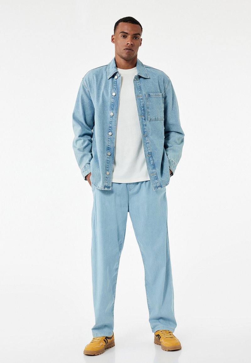 Lichtblauwe denimjack met knopen, wit T-shirt eronder, lichtblauwe broek en gele sneakers. Gladde textuur en relaxte pasvorm.