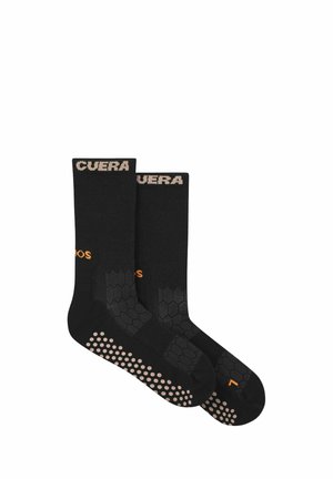 Paire de chaussettes noires mi-mollet avec des points antidérapants beiges sur les semelles, motif hexagonal sur le pied et texte beige sur le bord supérieur.