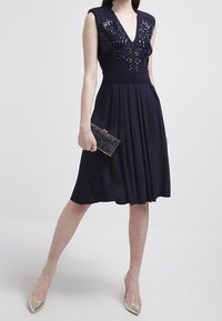 Marinblå ärmlös klänning med V-ringning och blommönsterdekorationer vid bröstet. Utsvängd kjol, kombinerad med en glittrande kuvertväska och metallfärgade klackar.