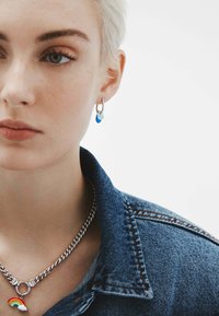 Pendiente de aro plateado con encantos en forma de corazón azul y azul claro. La modelo lleva un collar de cadena plateado con un pequeño colgante de arcoíris. Se ve una chaqueta de mezclilla.