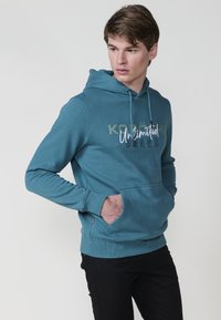 Felpa turchese con tasca a kangaroo, cappuccio con cordino e testo stampato "KOÜCH" e "Unlimited Brand". Tessuto morbido, vestibilità casual.