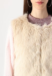 Gilet en fausse fourrure beige avec une surface douce et texturée, porté sur un pull en tricot rayé rose et gris. Grandes boucles d'oreilles rondes visibles.
