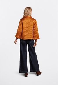 Kobieta odwrócona tyłem, ubrana w pikowaną pomarańczową kurtkę, szerokie ciemne dżinsy, brązowe botki na obcasie i pasiastą koszulę z długim rękawem pod spodem.