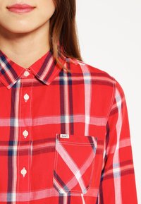 Chemise à carreaux rouges en coton avec un col boutonné, présentant des motifs de grille blancs et bleus, et une poche avant avec une étiquette.