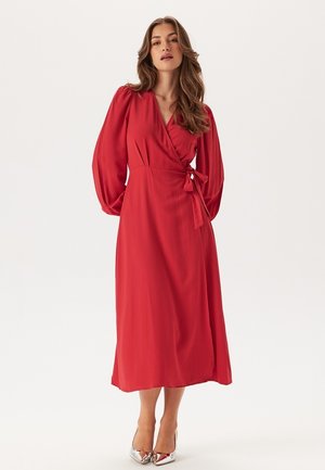 Abito avvolgente rosso con maniche lunghe a palloncino, vita legata e gonna fluida di lunghezza media. Tessuto morbido, colore uniforme e décolleté argentati.