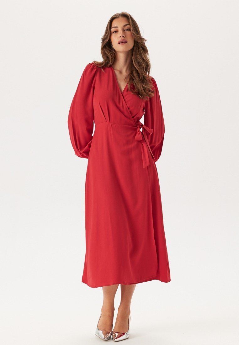 Robe rouge à manches bouffantes longues, avec un détail de taille nouée et une jupe fluide mi-longue. Tissu lisse, couleur unie et talons argentés.