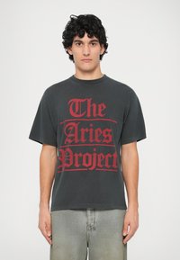 Siva oversized majica od pamuka, s velikim crvenim grafičkim natpisom "The Aries Project" otisnutim na prednjem dijelu.