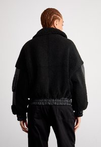 Zwarte fleecejas met een pluizige textuur, oversized kraag, elastische zoom en contrasterende zwarte leren mouwen met een utility zak.