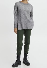 Grau gestrickter Pullover mit geripptem Saum, kombiniert mit grünen, schmalen Hosen. Das Model trägt schwarze Schnürstiefel im Combat-Stil. Einfaches Design, weicher Stoff.